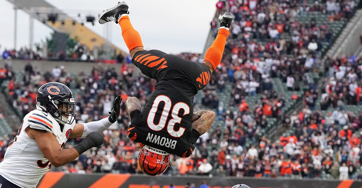 CINCINNATI, OHIO - NOVEMBER 02: Chase Brown #30 of the Cincinnati Bengals leaps over Kevin Byard III #31 of the Chicago Bears during the fourth quarter in the game at Paycor Stadium on November 02, 2025 in Cincinnati, Ohio. Brown is completely upside down, back to the camera, legs in the air, looking like his flip will continue to land him on his back. Both Bears players are looking at him. One is on the ground, behind him, looking up. The other is reaching toward Brown. In the background is a full stadium of people, and from the orange it's clear it's the Bengals stadium.