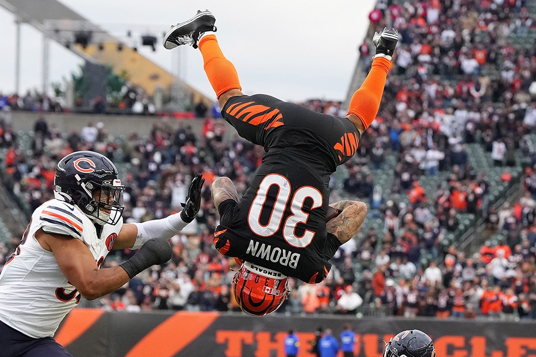 CINCINNATI, OHIO - NOVEMBER 02: Chase Brown #30 of the Cincinnati Bengals leaps over Kevin Byard III #31 of the Chicago Bears during the fourth quarter in the game at Paycor Stadium on November 02, 2025 in Cincinnati, Ohio. Brown is completely upside down, back to the camera, legs in the air, looking like his flip will continue to land him on his back. Both Bears players are looking at him. One is on the ground, behind him, looking up. The other is reaching toward Brown. In the background is a full stadium of people, and from the orange it's clear it's the Bengals stadium.