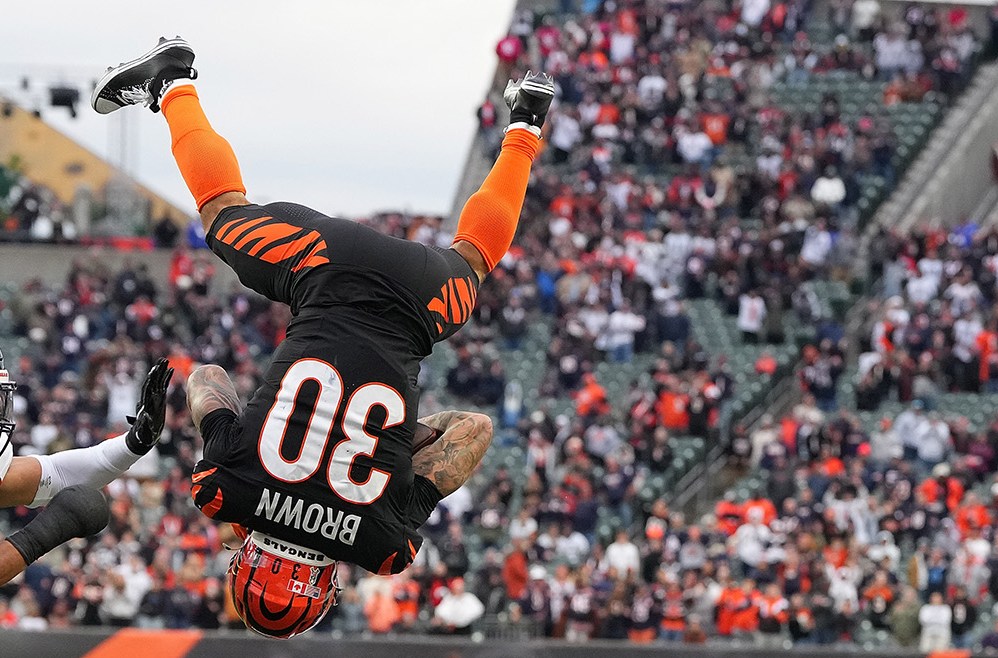 CINCINNATI, OHIO - NOVEMBER 02: Chase Brown #30 of the Cincinnati Bengals leaps over Kevin Byard III #31 of the Chicago Bears during the fourth quarter in the game at Paycor Stadium on November 02, 2025 in Cincinnati, Ohio. Brown is completely upside down, back to the camera, legs in the air, looking like his flip will continue to land him on his back. Both Bears players are looking at him. One is on the ground, behind him, looking up. The other is reaching toward Brown. In the background is a full stadium of people, and from the orange it's clear it's the Bengals stadium.