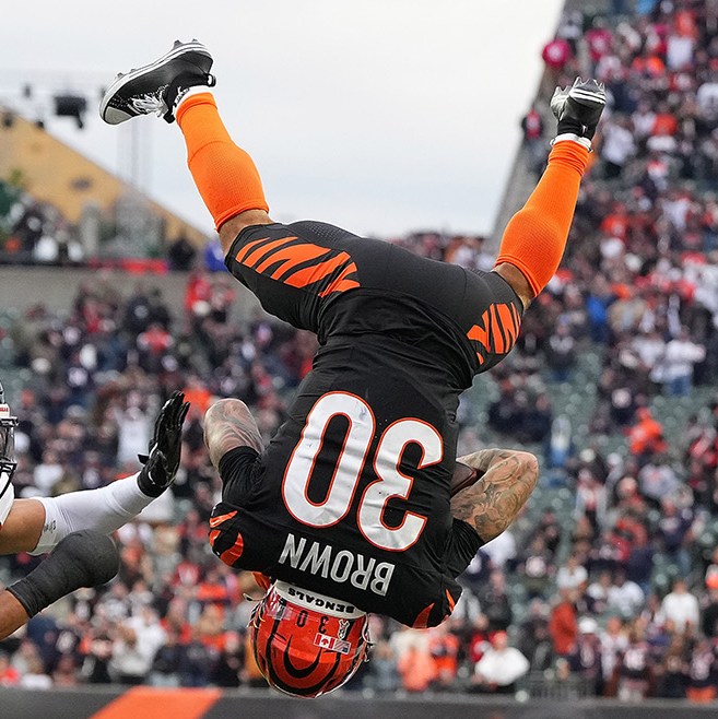 CINCINNATI, OHIO - NOVEMBER 02: Chase Brown #30 of the Cincinnati Bengals leaps over Kevin Byard III #31 of the Chicago Bears during the fourth quarter in the game at Paycor Stadium on November 02, 2025 in Cincinnati, Ohio. Brown is completely upside down, back to the camera, legs in the air, looking like his flip will continue to land him on his back. Both Bears players are looking at him. One is on the ground, behind him, looking up. The other is reaching toward Brown. In the background is a full stadium of people, and from the orange it's clear it's the Bengals stadium.