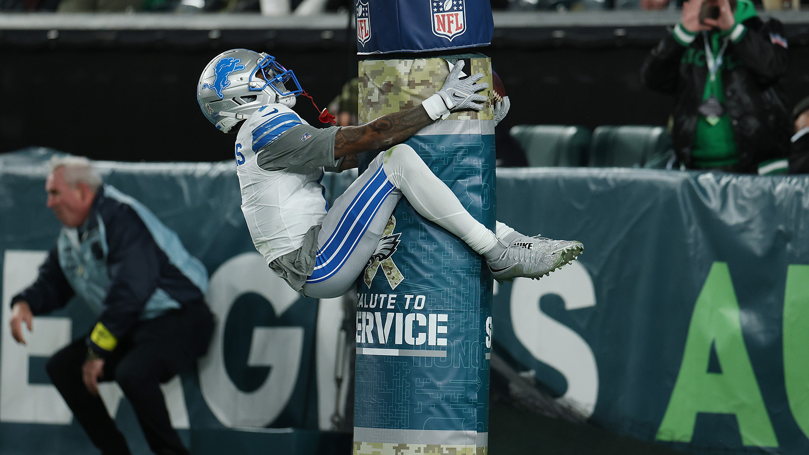 PHILADELPHIA, PENNSYLVANIA - NOVEMBER 16: Jameson Williams #1 of the Detroit Lions celebrates a touchdown during the first half against the Philadelphia Eagles at Lincoln Financial Field on November 16, 2025 in Philadelphia, Pennsylvania. Williams is hugging the goal post after scoring a touchdown. He's in his white and silver and blue Lions jersey and he's pretty high up on the goalpost, above the NFL "Salute to Service" logo and he's clearly very excited.