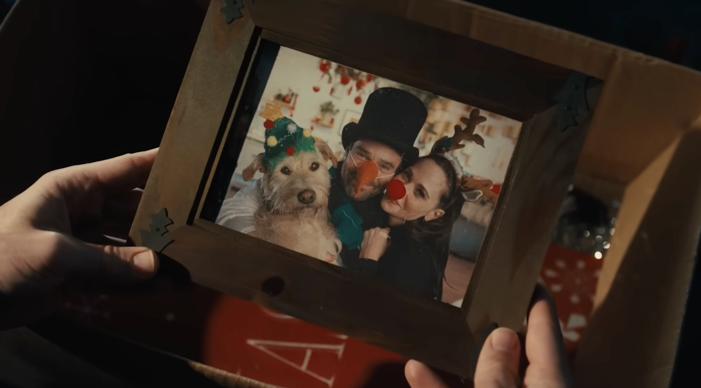 a hand holds a framed photo of Zooey Deschanel, Charlie Cox, and a terrier mix played by Gus the Dog. Everyone is in christmas attire