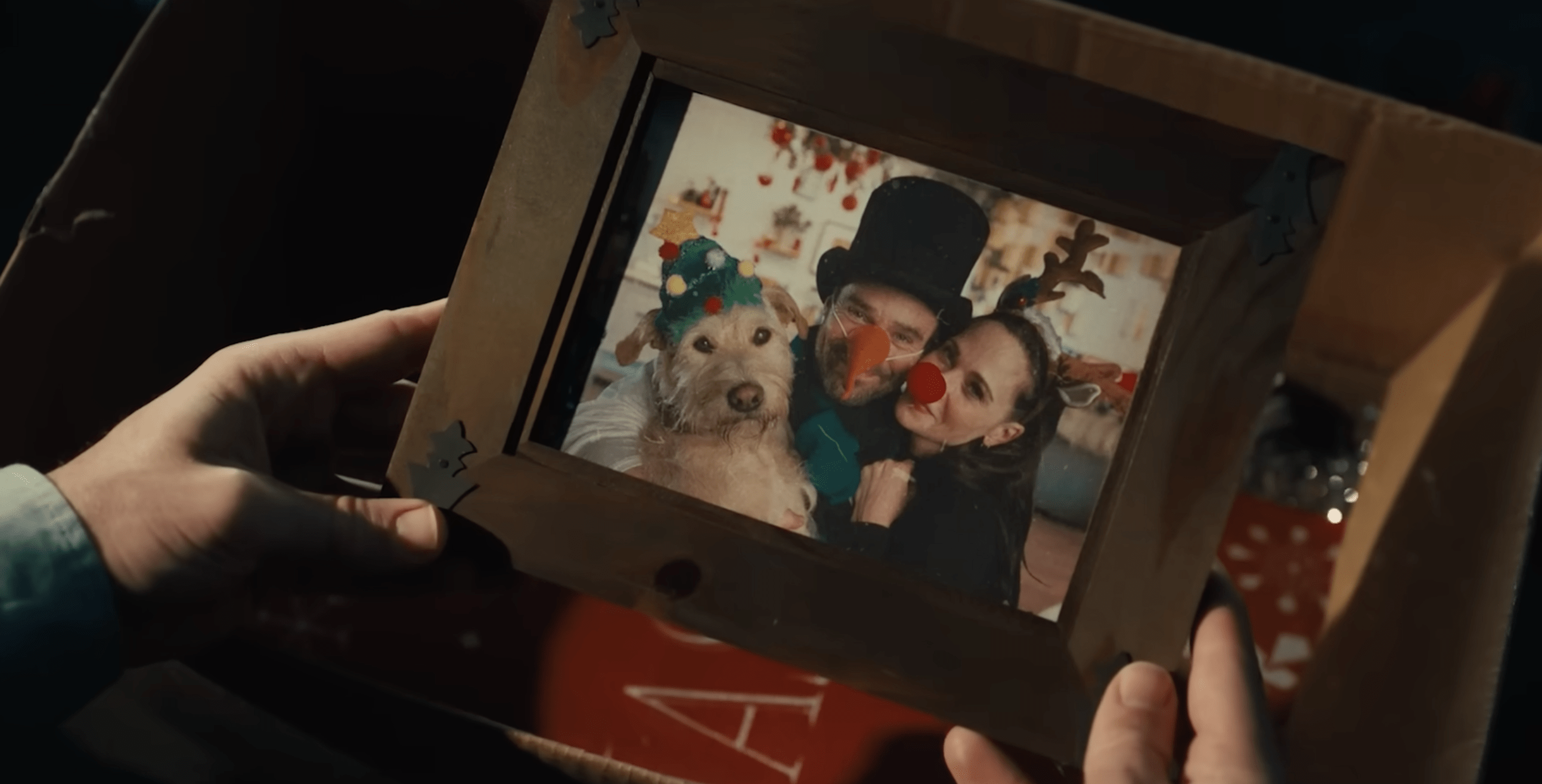a hand holds a framed photo of Zooey Deschanel, Charlie Cox, and a terrier mix played by Gus the Dog. Everyone is in christmas attire