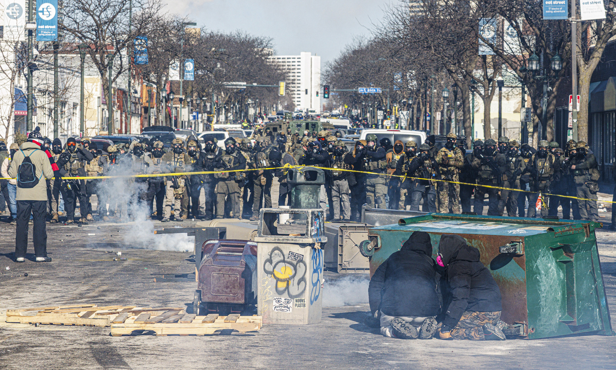 Protesters take cover behind overturned trash containers as a line of federal agents forms a barricade across a downtown street during clashes following the fatal shooting of a demonstrator earlier in the day, on January 24, 2026 in Minneapolis, Minnesota.