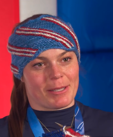 A still image of Breezy Johnson in the weird headband she knitted for herself. In the foreground, her knitting needles are visible.
