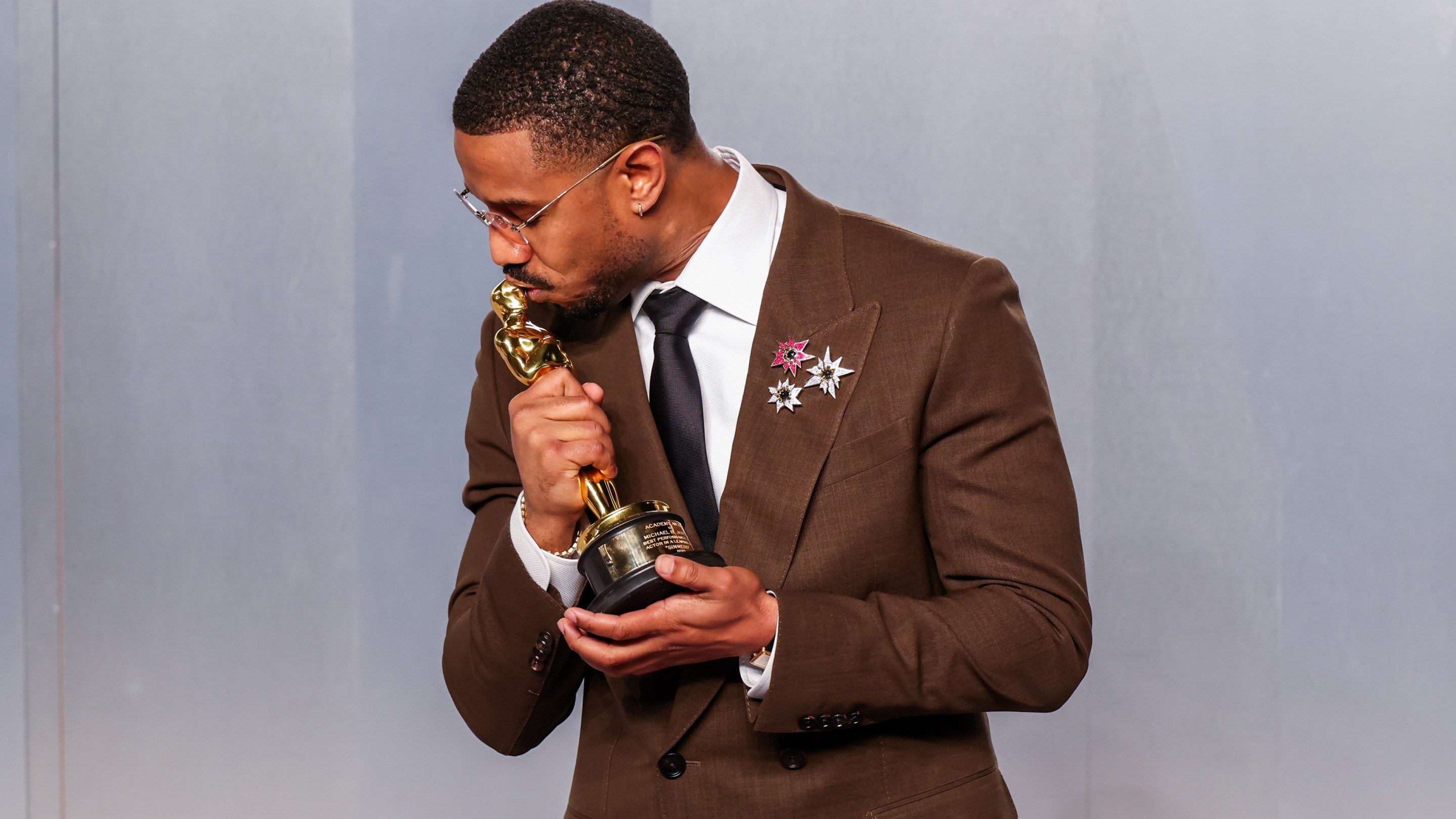 Michael B. Jordan kisses his oscar wearing a brown suit with three small broaches on the lapel.