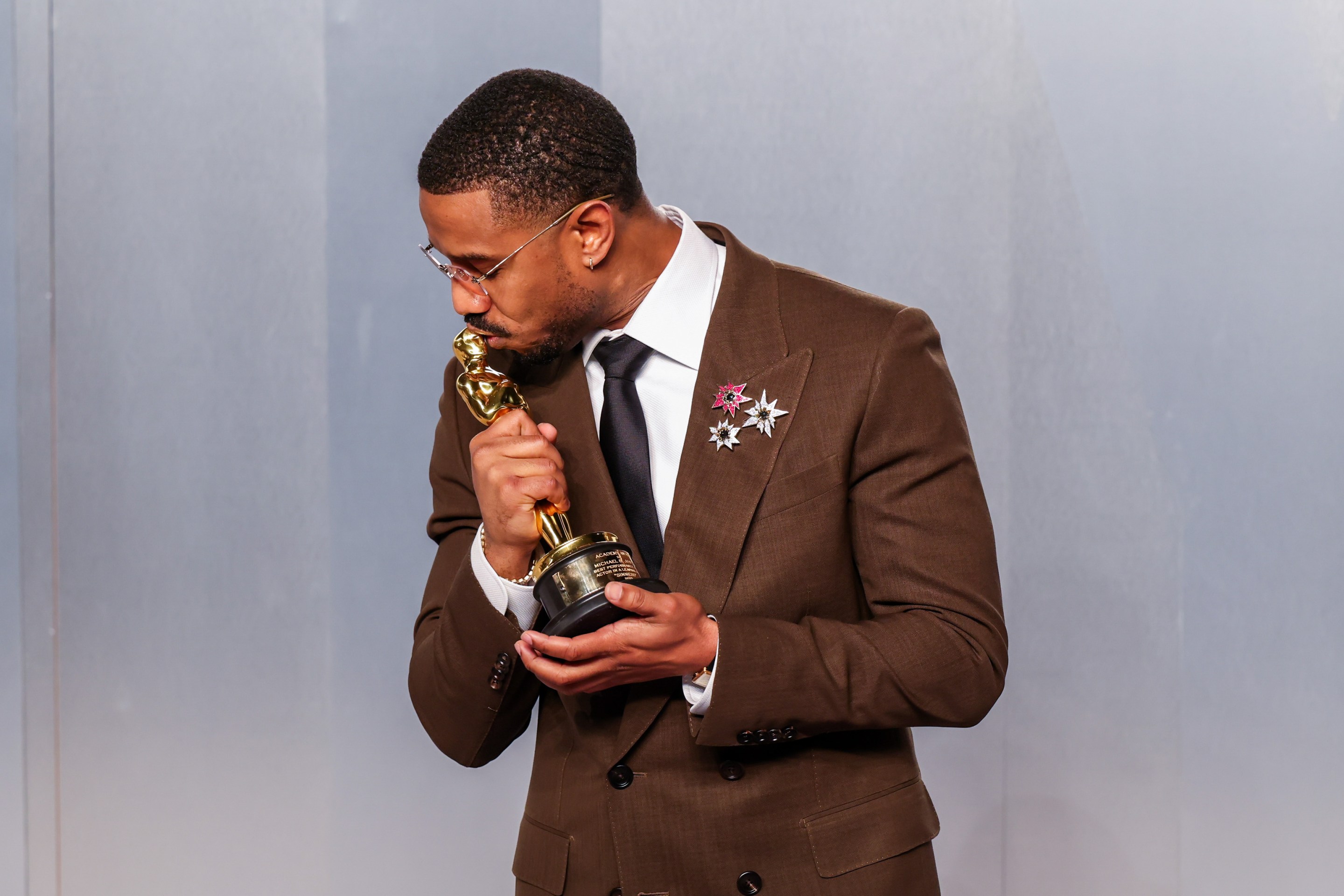 Michael B. Jordan kisses his oscar wearing a brown suit with three small broaches on the lapel.