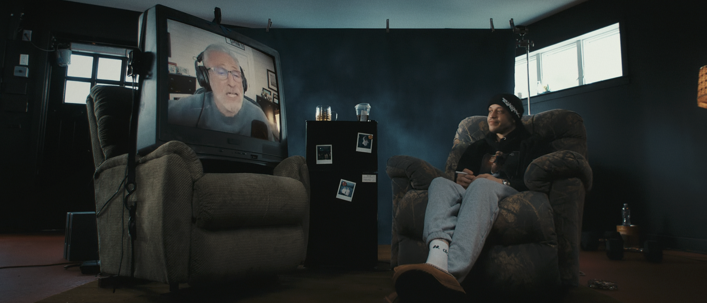 Pete Davidson sits in a garage on an armchair. There's a giant TV on a second armchair with an image of Jon Stewart calling in remotely,