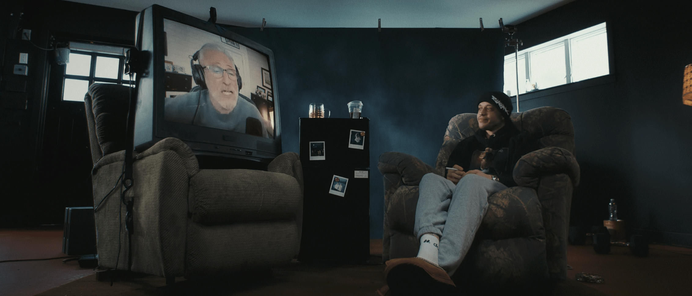 Pete Davidson sits in a garage on an armchair. There's a giant TV on a second armchair with an image of Jon Stewart calling in remotely,