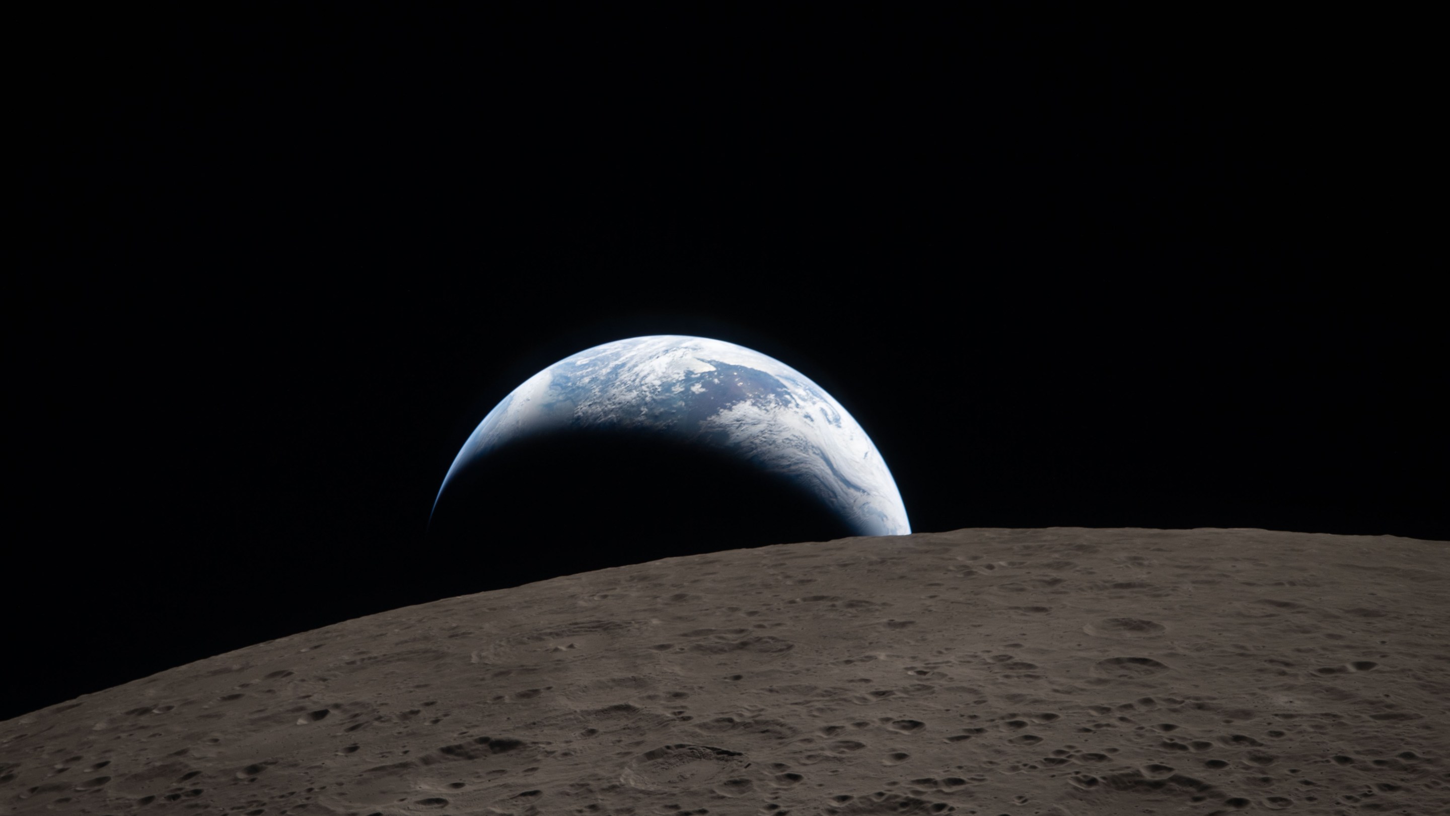 Earthset captured through the Orion spacecraft window at 6:41 p.m. EDT, April 6, 2026, during the Artemis II crew’s flyby of the Moon. A muted blue Earth with bright white clouds sets behind the cratered lunar surface. The dark portion of Earth is experiencing nighttime. On Earth’s day side, swirling clouds are visible over the Australia and Oceania region. In the foreground, Ohm crater has terraced edges and a flat floor interrupted by central peaks. Central peaks form in complex craters when the lunar surface, liquefied on impact, splashes upwards during the crater’s formation.
