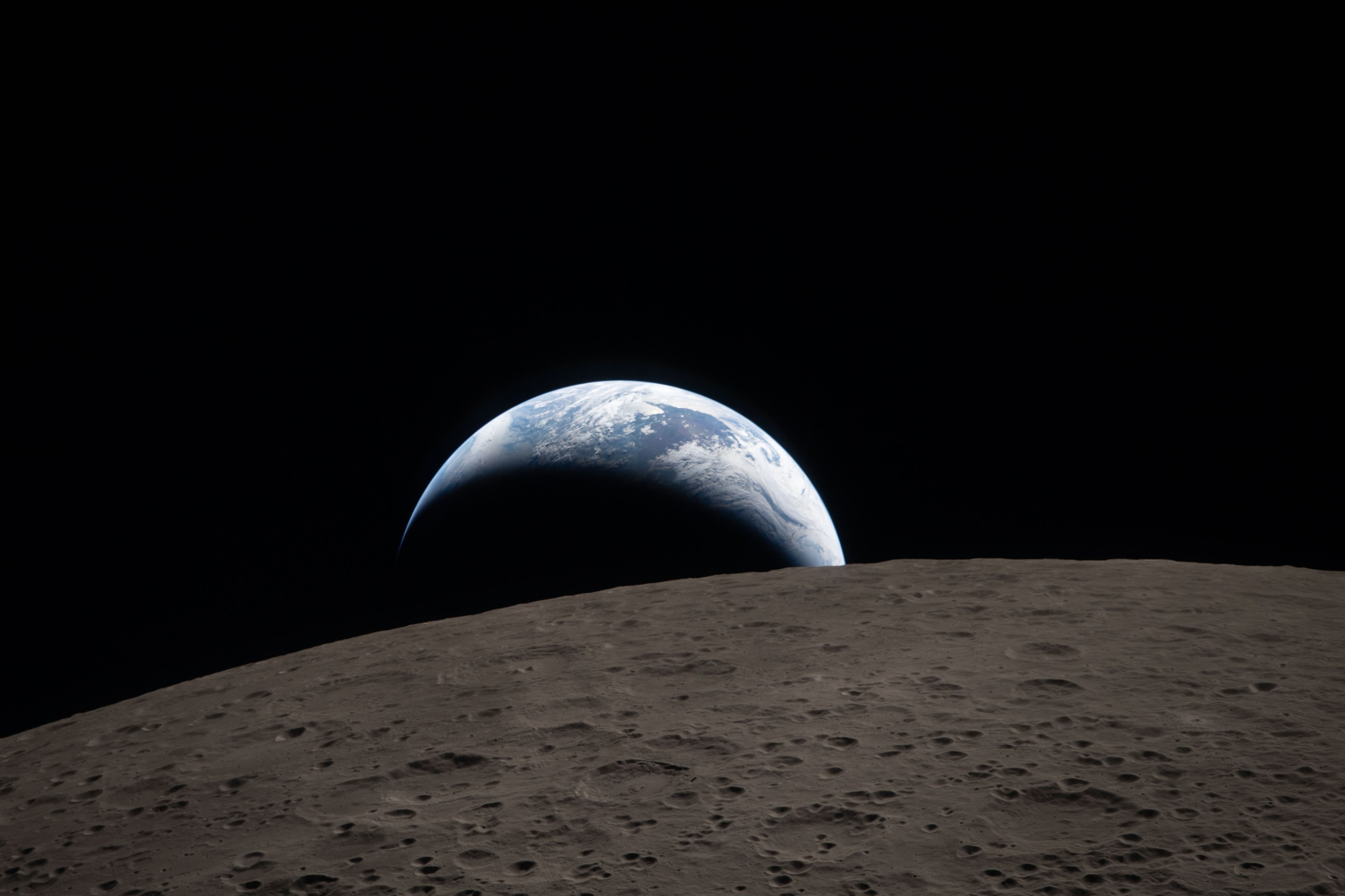 Earthset captured through the Orion spacecraft window at 6:41 p.m. EDT, April 6, 2026, during the Artemis II crew’s flyby of the Moon. A muted blue Earth with bright white clouds sets behind the cratered lunar surface. The dark portion of Earth is experiencing nighttime. On Earth’s day side, swirling clouds are visible over the Australia and Oceania region. In the foreground, Ohm crater has terraced edges and a flat floor interrupted by central peaks. Central peaks form in complex craters when the lunar surface, liquefied on impact, splashes upwards during the crater’s formation.