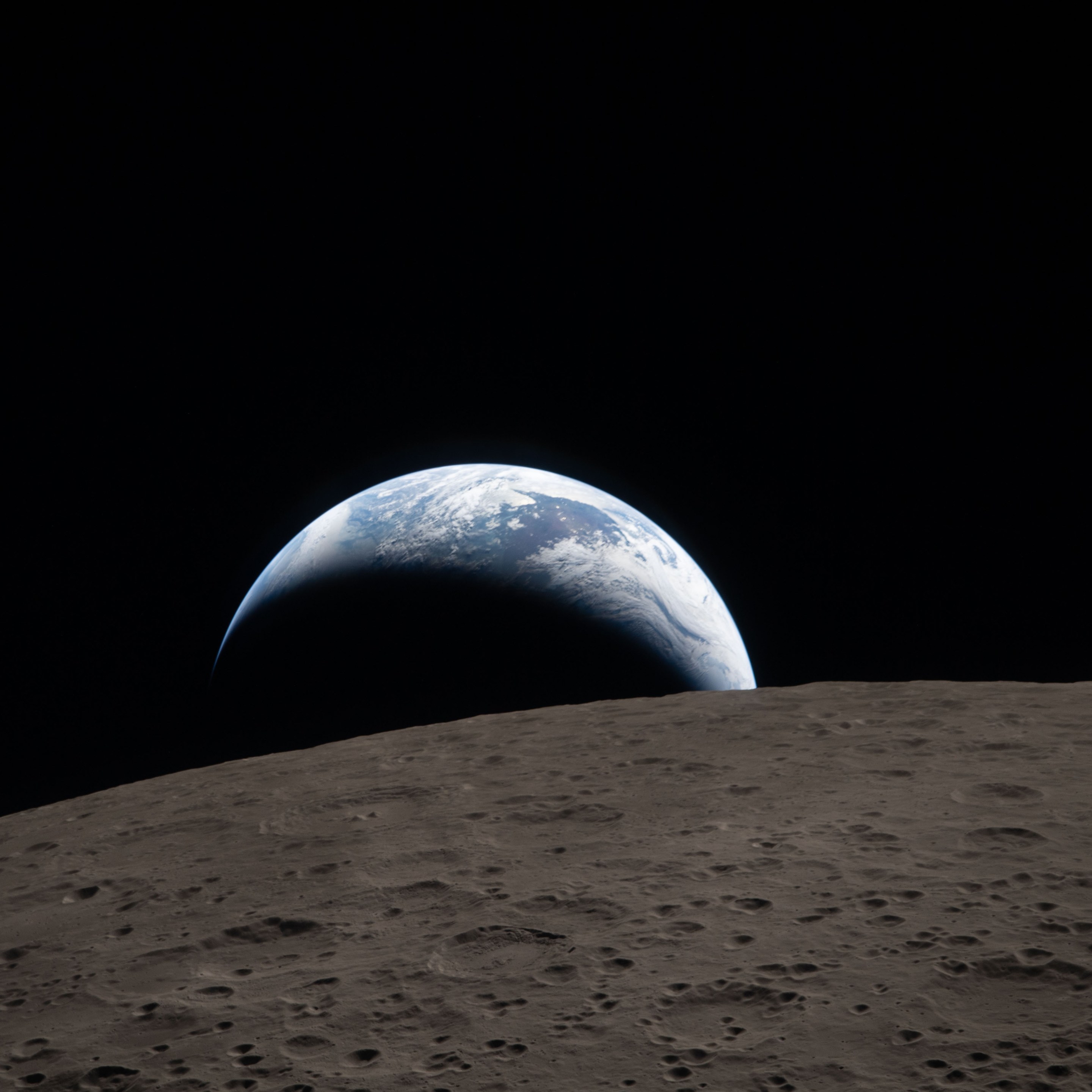 Earthset captured through the Orion spacecraft window at 6:41 p.m. EDT, April 6, 2026, during the Artemis II crew’s flyby of the Moon. A muted blue Earth with bright white clouds sets behind the cratered lunar surface. The dark portion of Earth is experiencing nighttime. On Earth’s day side, swirling clouds are visible over the Australia and Oceania region. In the foreground, Ohm crater has terraced edges and a flat floor interrupted by central peaks. Central peaks form in complex craters when the lunar surface, liquefied on impact, splashes upwards during the crater’s formation.