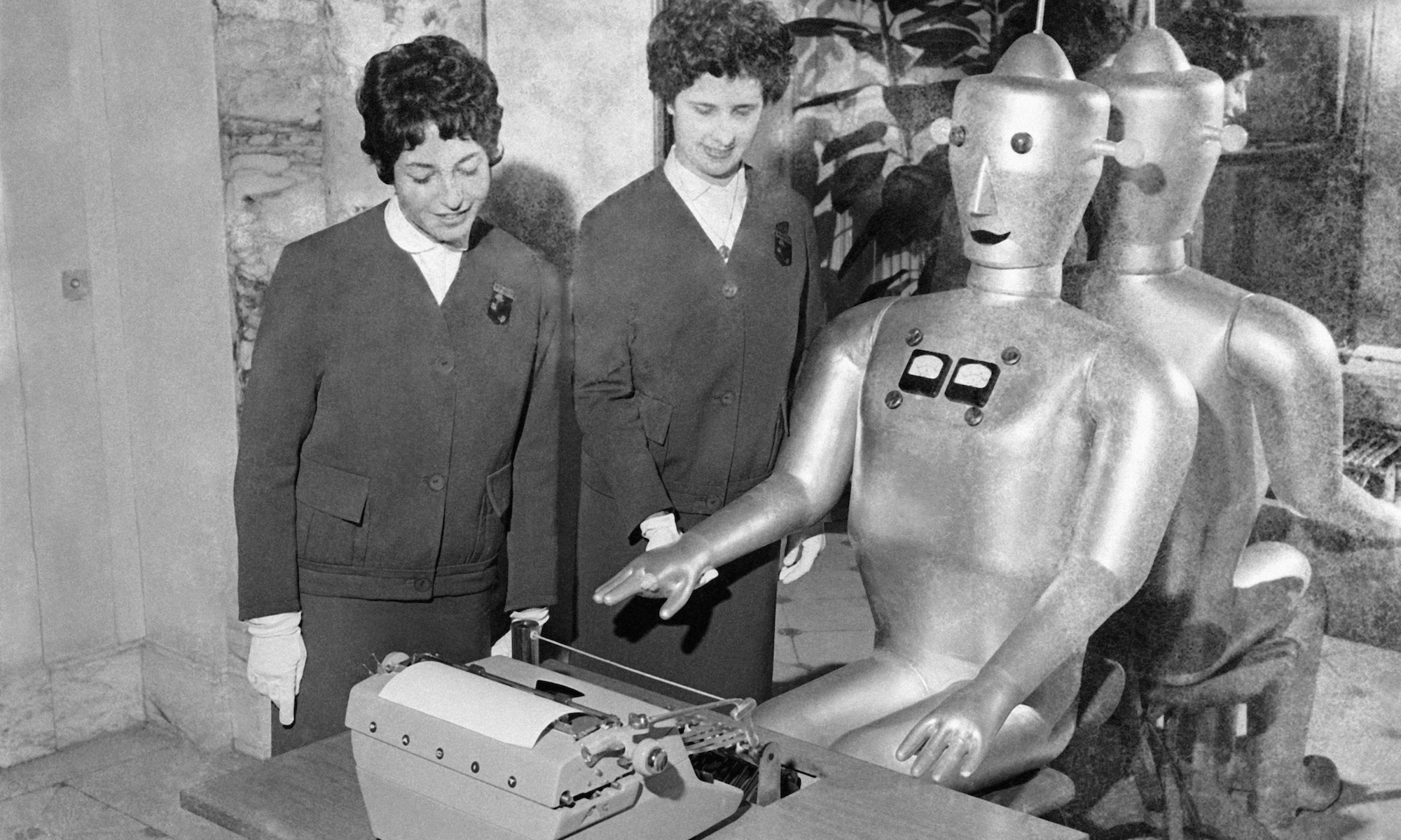 In a black-and-white old photo from an exhibition in Paris in 1960, a robotic typist sits at a desk in front of a typewriter, with two women looking on.