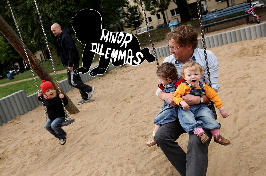 A father with twins sits on a swing in a playground while a another child swings alone nearby.