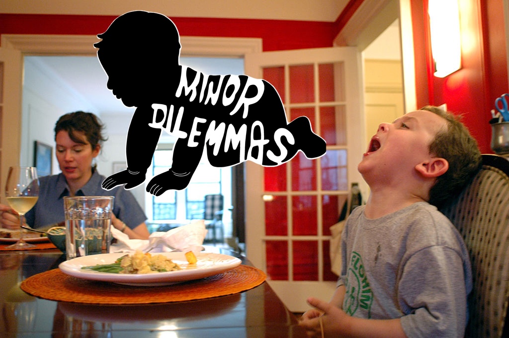 A woman sits at the dinner table eating with her young son, who is making a funny face while his plate of food sits untouched. The black and white minor dilemmas logo is in the middle of the image.