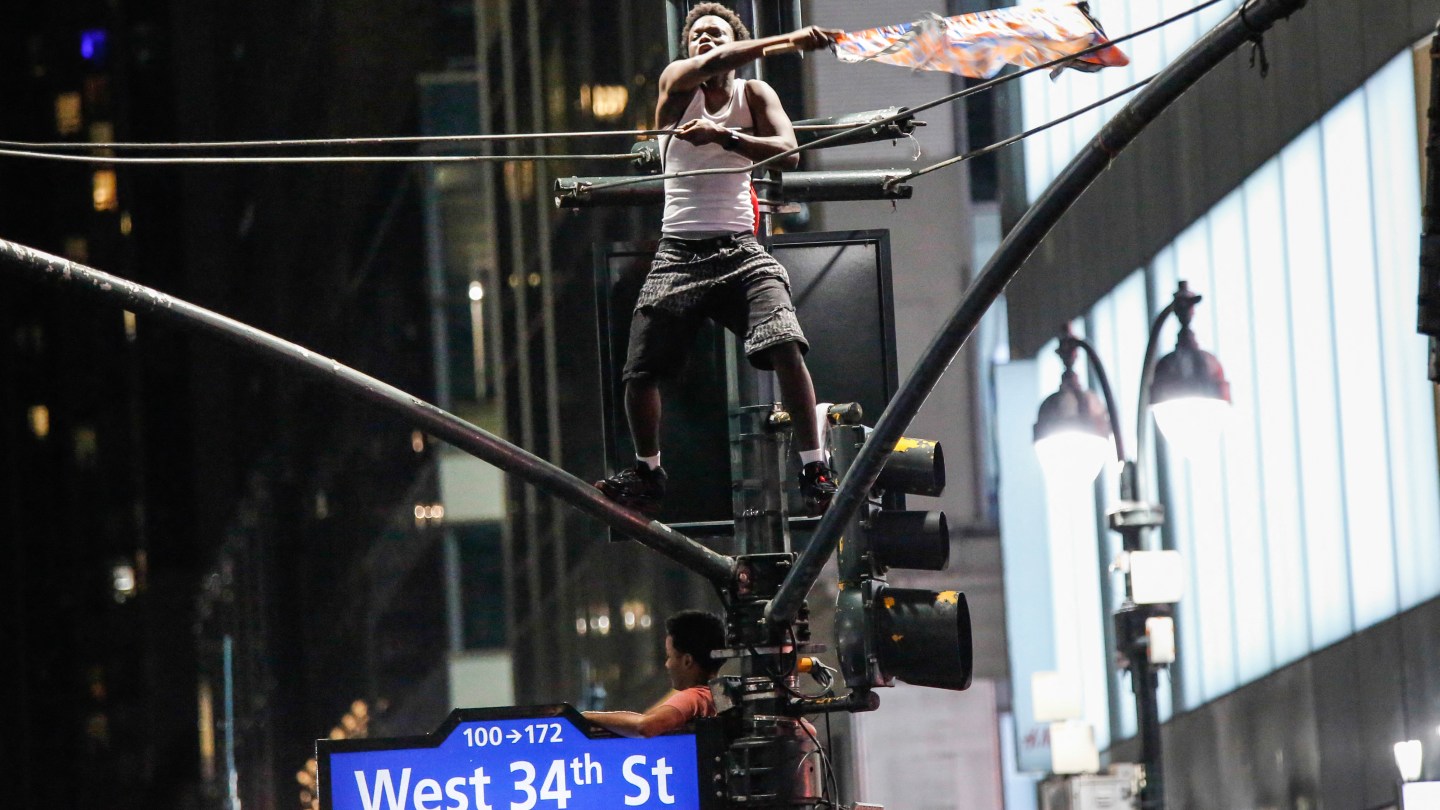 A Knicks fan waves a flag while standing on top of a traffic light poles after the New York Knicks defeated the San Antonio Spurs in Game 5 of the NBA Finals to win the series, in New York, on June 14, 2026. The Knicks won the NBA Championship for the first time since 1973, for the third time in franchise history. (Photo by kena betancur / AFP via Getty Images)