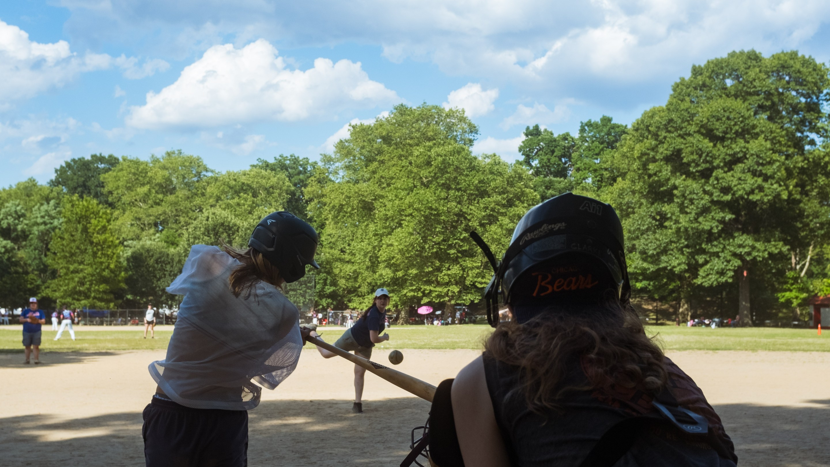 view from behind the catcher on a park ballfield as a pitch comes in and a hitter swings