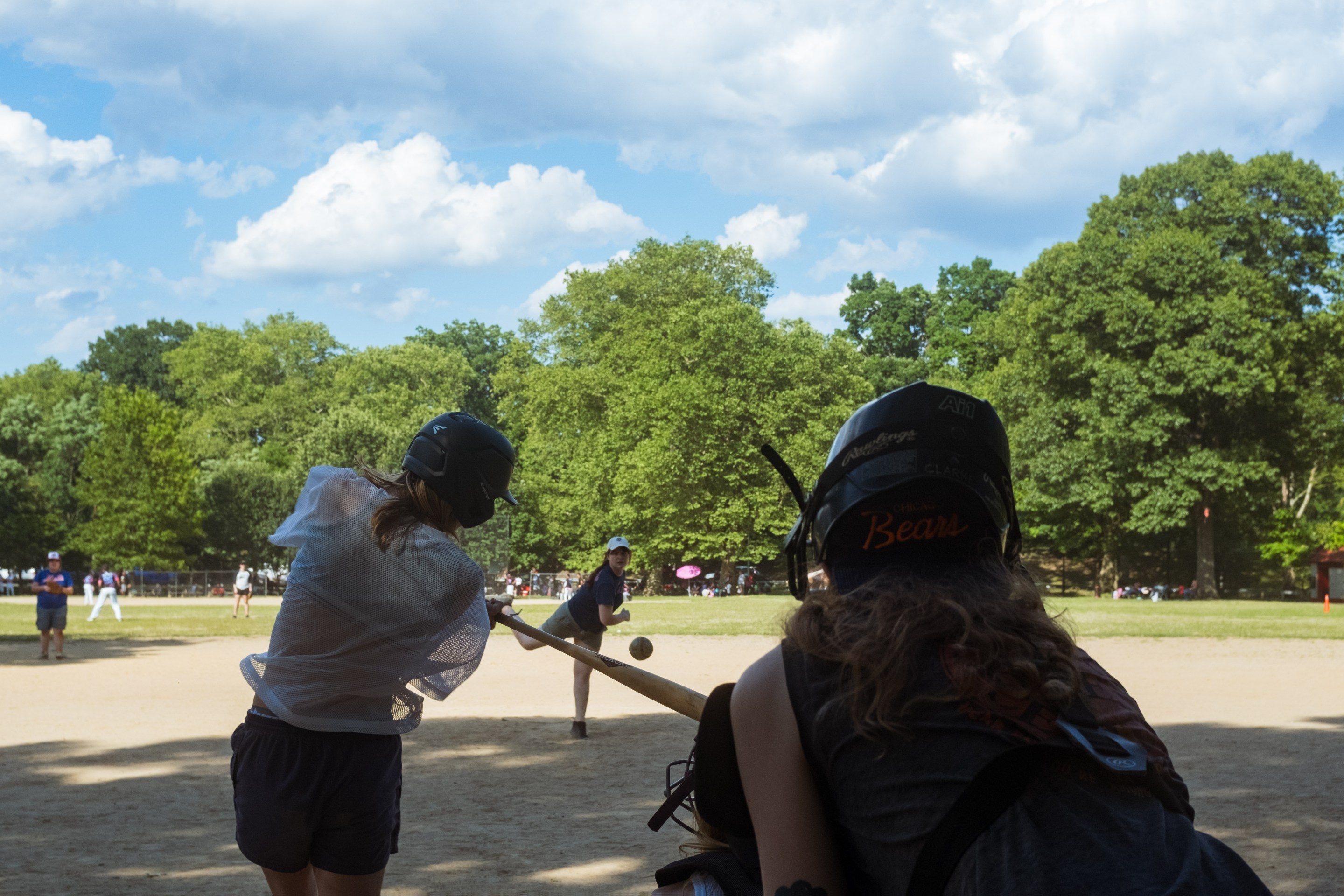 view from behind the catcher on a park ballfield as a pitch comes in and a hitter swings