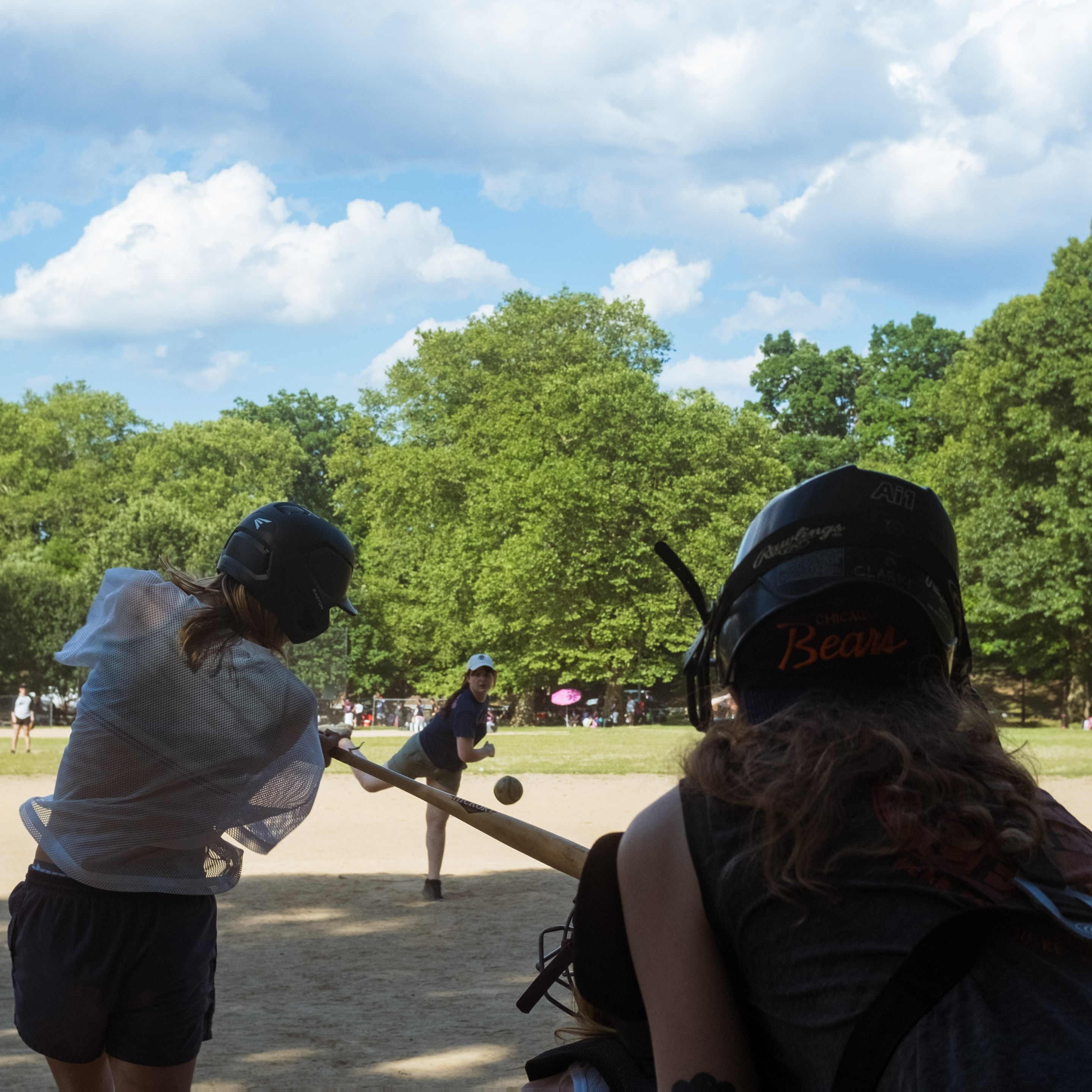 view from behind the catcher on a park ballfield as a pitch comes in and a hitter swings