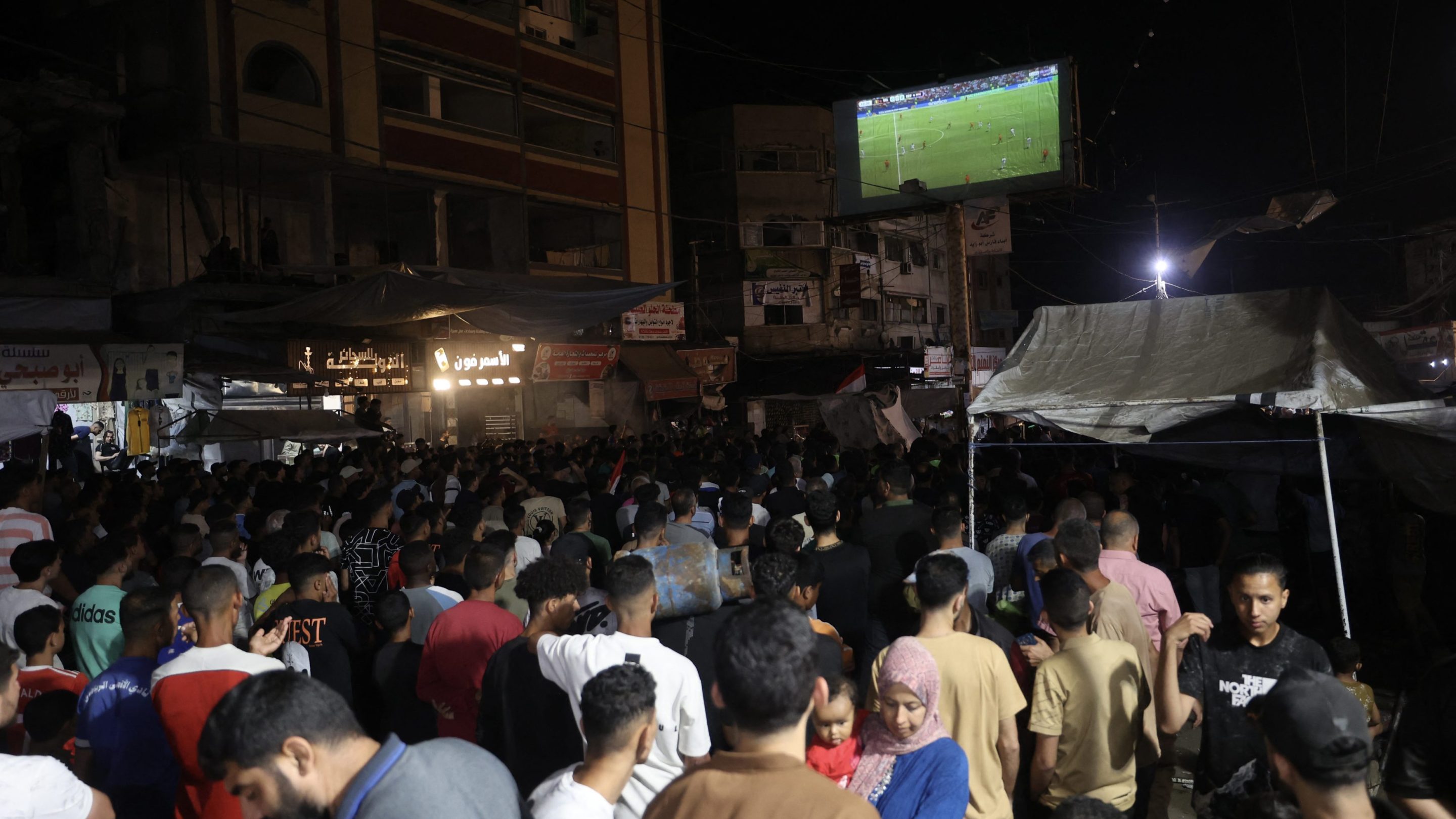 Palestinian football fans displaced from their homes, which were destroyed by Israeli military strikes, watch Belgium vs. Egypt on a giant screen at the Nuseirat refugee camp in Gaza.