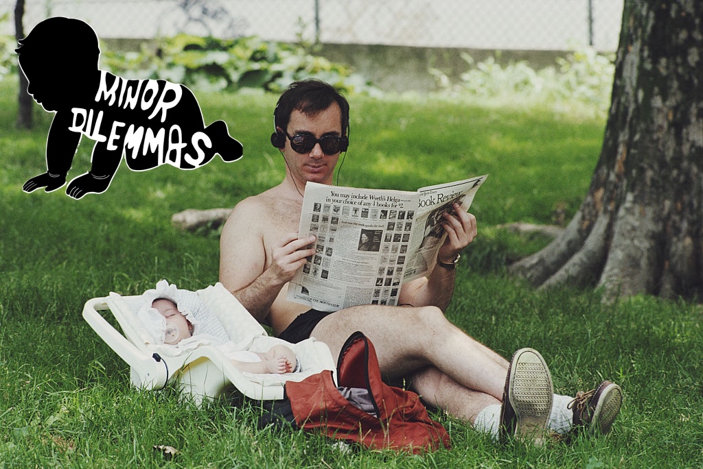 A dad sits in the grass at a park with his shirt off listening to music on headphones and reading the paper while his baby sleeps next to him