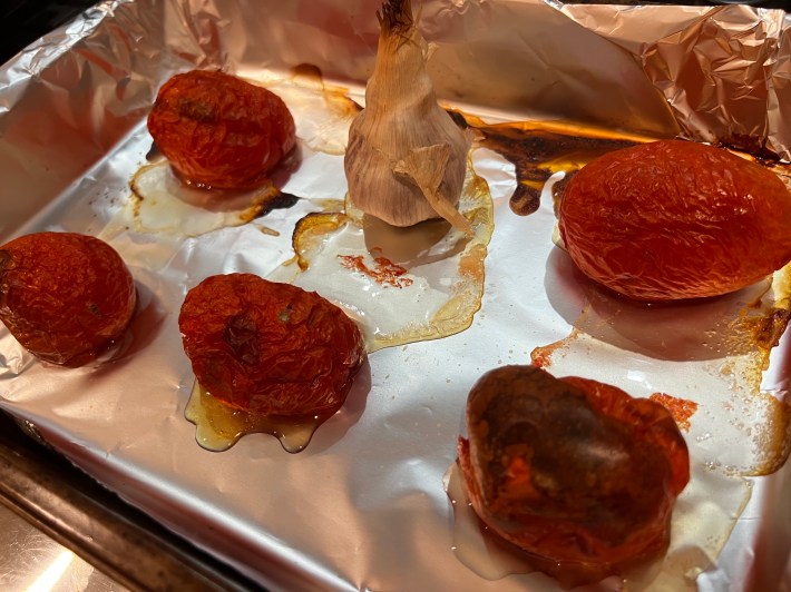 Five roasted Roma tomatoes and a head of garlic sit in a baking pan.