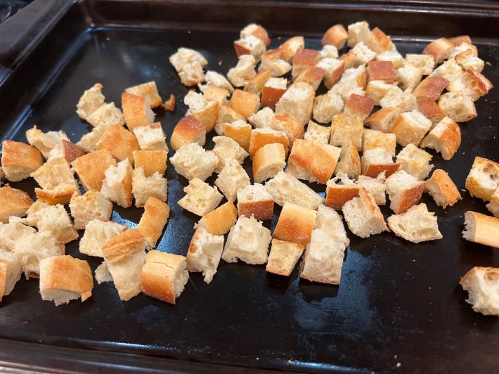 Lightly toasted cubes of bread are spread on a cookie sheet.