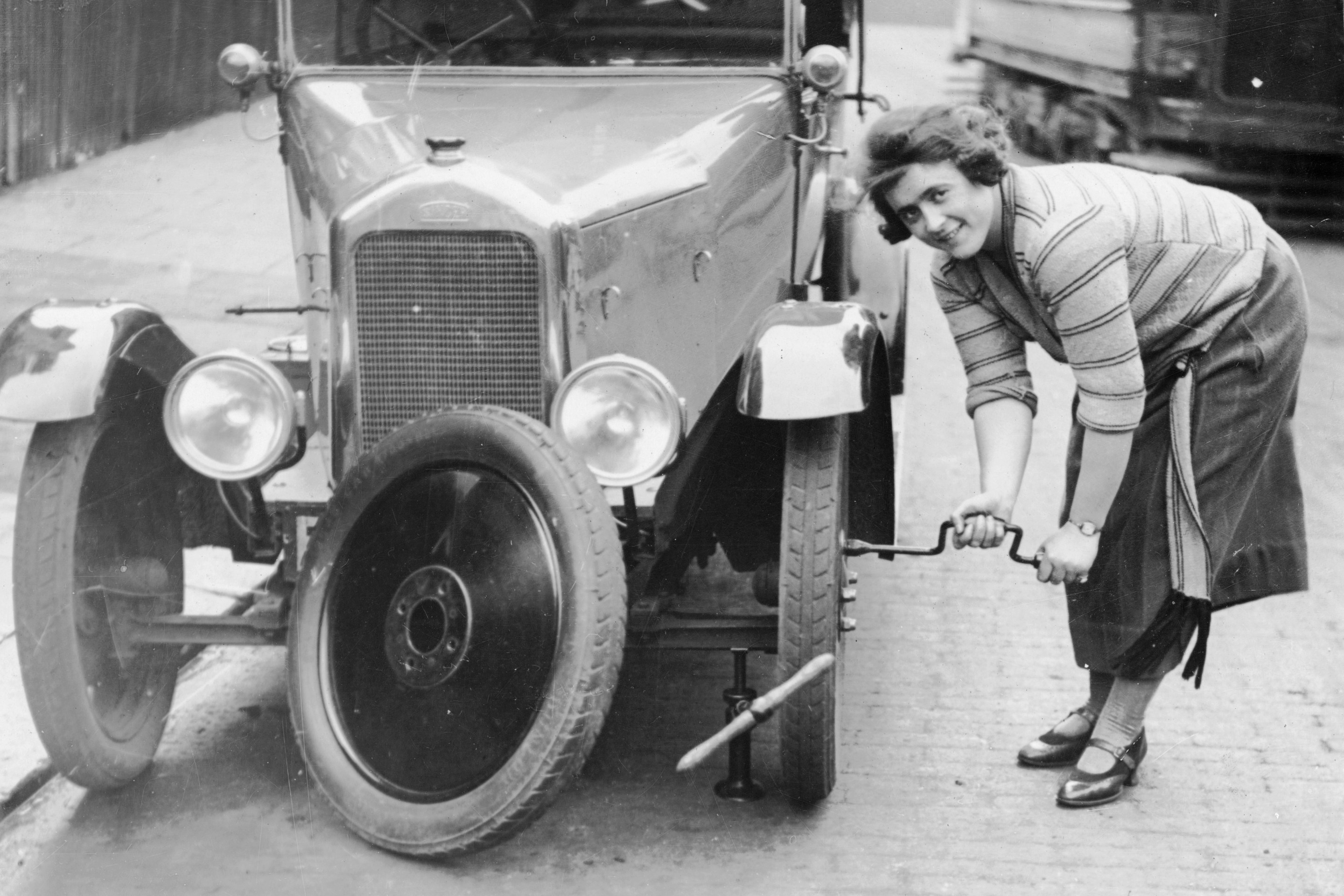Ivy Cummings changing a tyre on a 1925 Singer 10/26, London, c1925. The car is jacked up; Ms Cummings uses a spanner to tighten the bolts on the new wheel. Ivy Cummings was a racing driver who also ran a garage in Putney Bridge Road, London. She carried out all the repairs herself as well as selling second-hand cars. The Duke of York congratulated her for her many motor racing wins.
