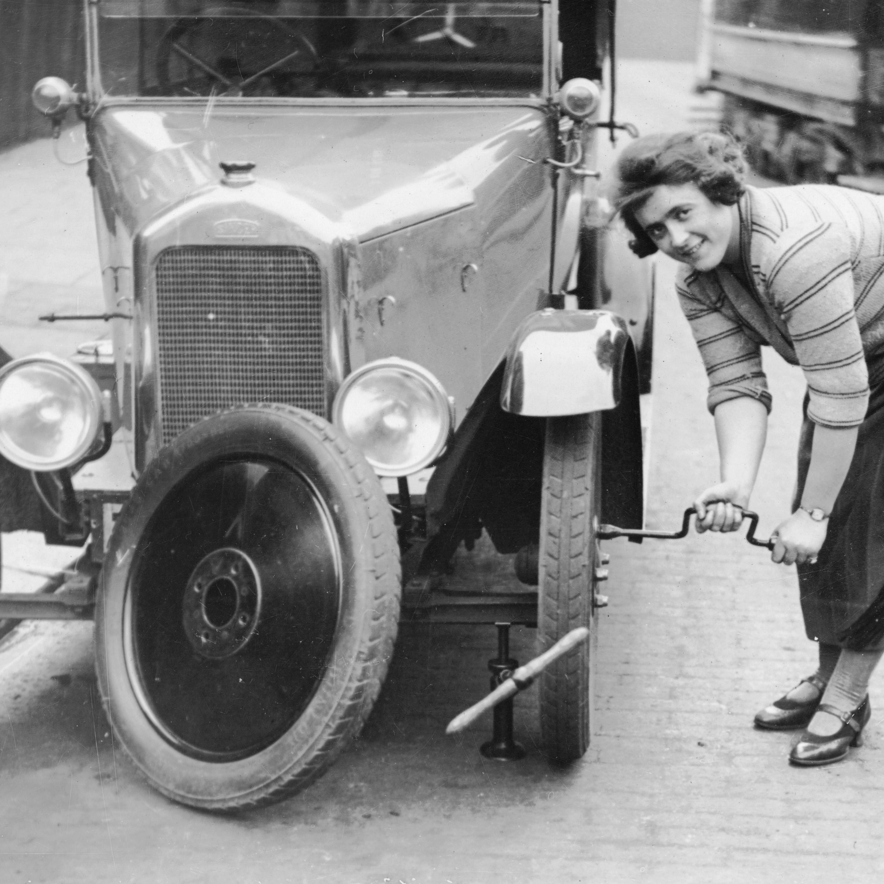 Ivy Cummings changing a tyre on a 1925 Singer 10/26, London, c1925. The car is jacked up; Ms Cummings uses a spanner to tighten the bolts on the new wheel. Ivy Cummings was a racing driver who also ran a garage in Putney Bridge Road, London. She carried out all the repairs herself as well as selling second-hand cars. The Duke of York congratulated her for her many motor racing wins.