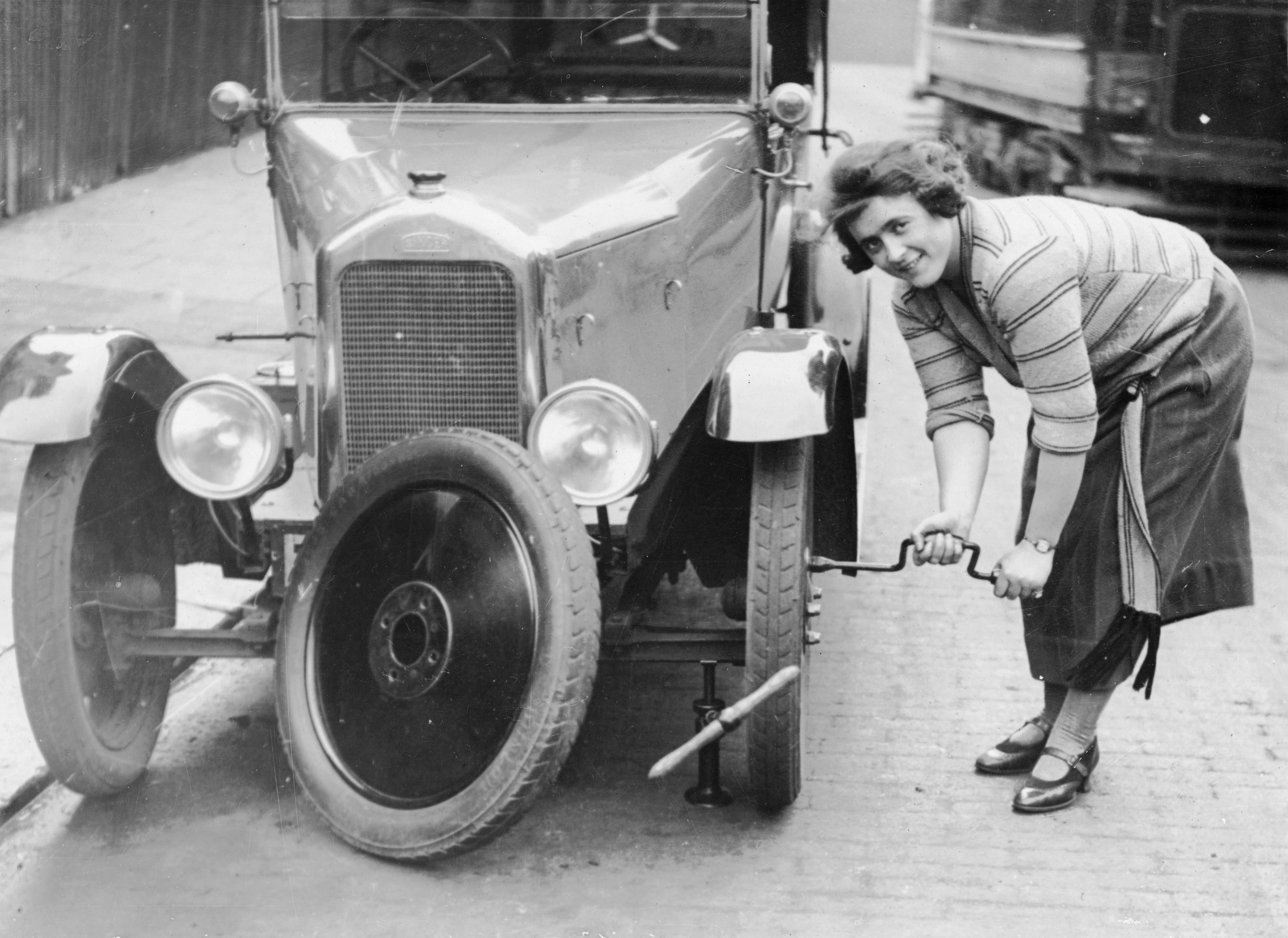 Ivy Cummings changing a tyre on a 1925 Singer 10/26, London, c1925. The car is jacked up; Ms Cummings uses a spanner to tighten the bolts on the new wheel. Ivy Cummings was a racing driver who also ran a garage in Putney Bridge Road, London. She carried out all the repairs herself as well as selling second-hand cars. The Duke of York congratulated her for her many motor racing wins.