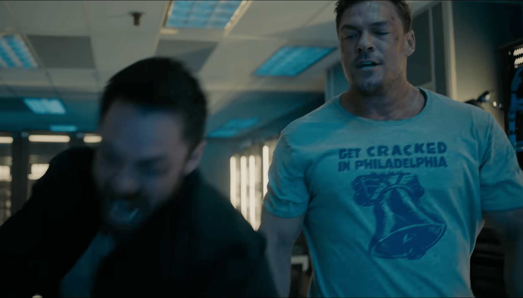 Alan Ritchson's Jack Reacher character glowers at a screaming man with his hand stuck in an industrial shredder during an episode from season four of Amazon's "Reacher." Reacher is wearing a t-shirt that says "Get Cracked In Philadelphia," and is huge.