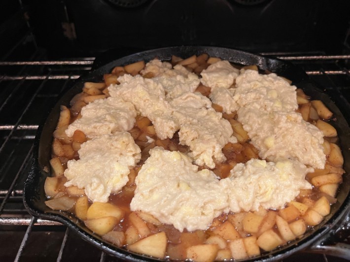 Raw biscuit dough on top of stewed apples, in a cast iron skillet, inside an oven.