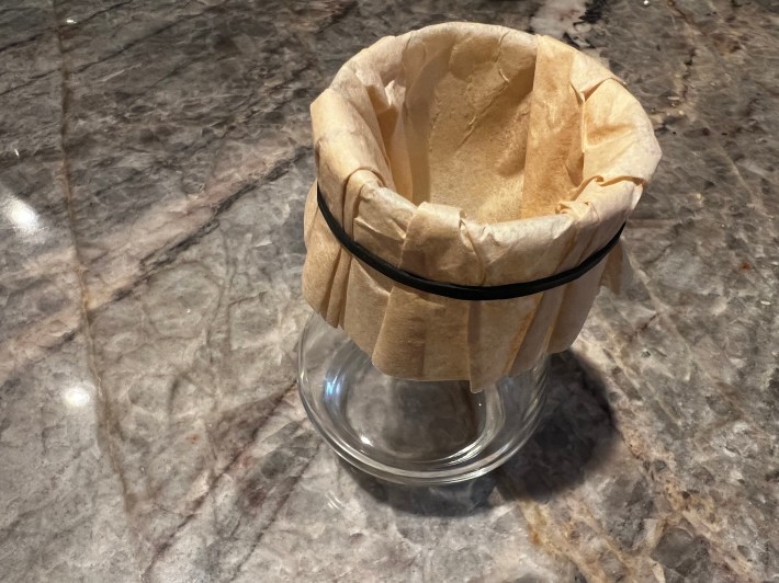 A coffee filter is wrapped around the rim of a whisky glass and secured by a rubber band.