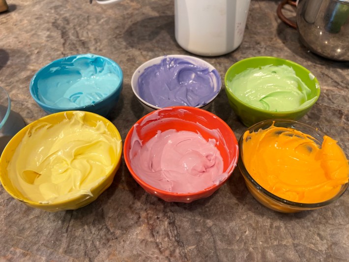 Six bowls containing dyed royal icing: blue, yellow, purple, pink, green, and orange.
