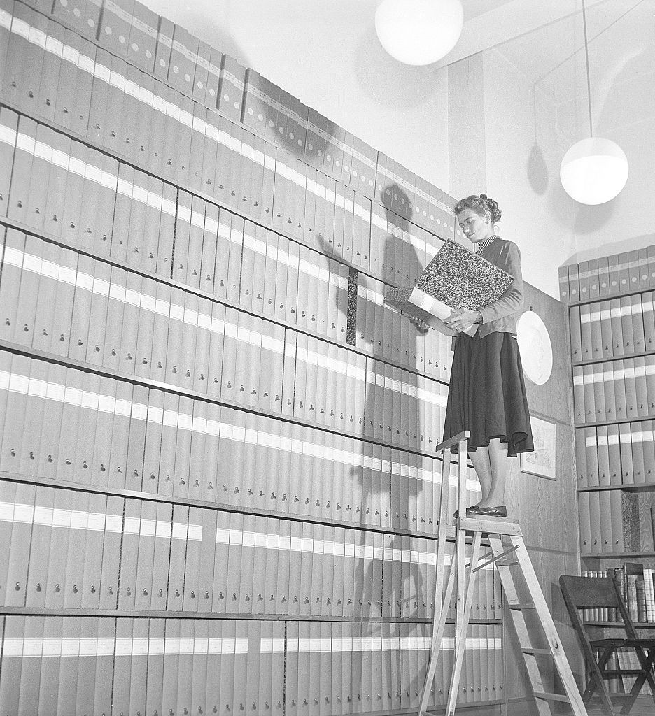 1950s woman at work. A young woman is standing on a ladder in an archive with shelfes filled with folders containing documents.