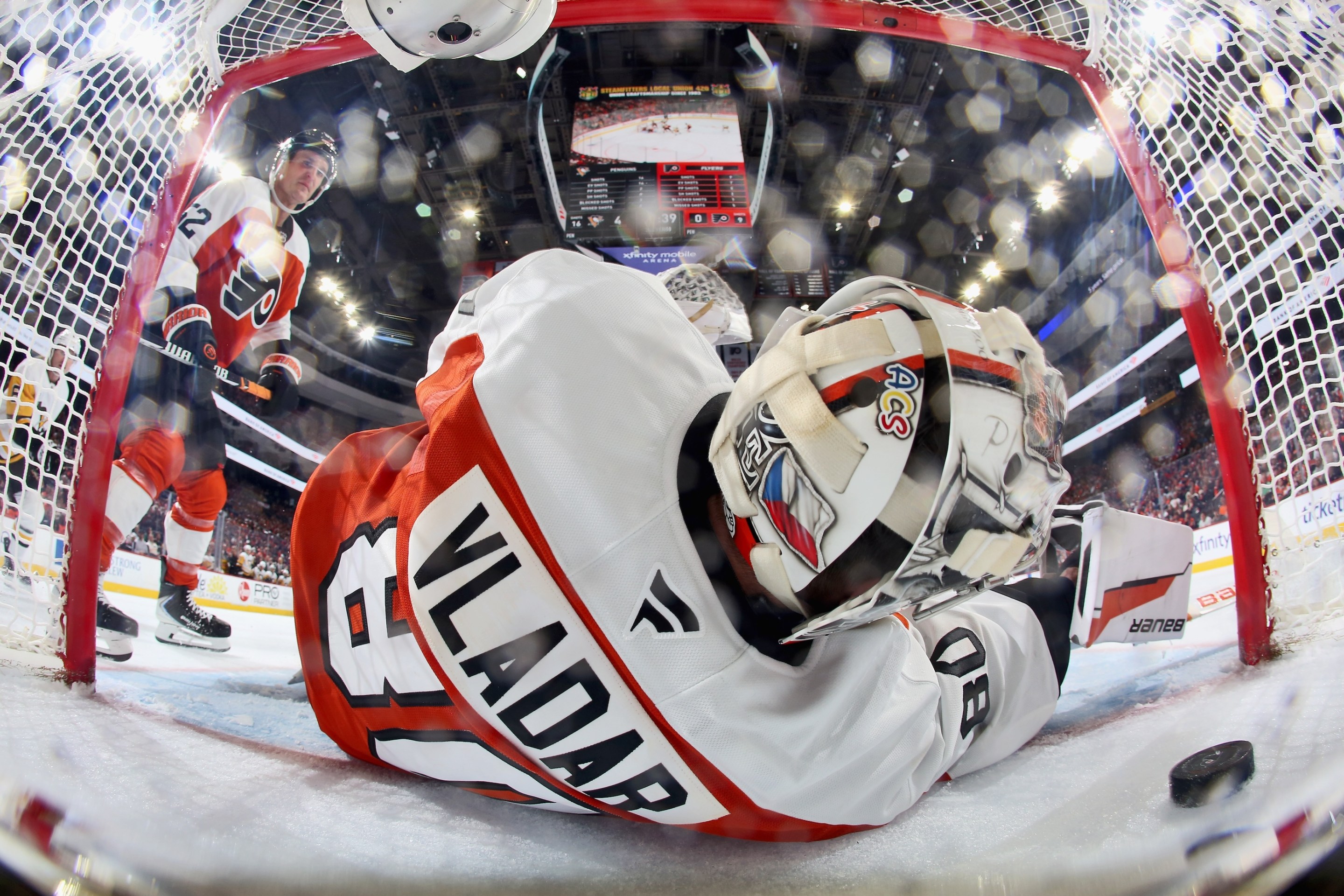 Dan Vladar of the Philadelphia Flyers surrenders a goal during the second period against the Pittsburgh Penguins as Christian Dvorak #22 looks on. Half his body is in the goal, and the puck is visible at lower right.