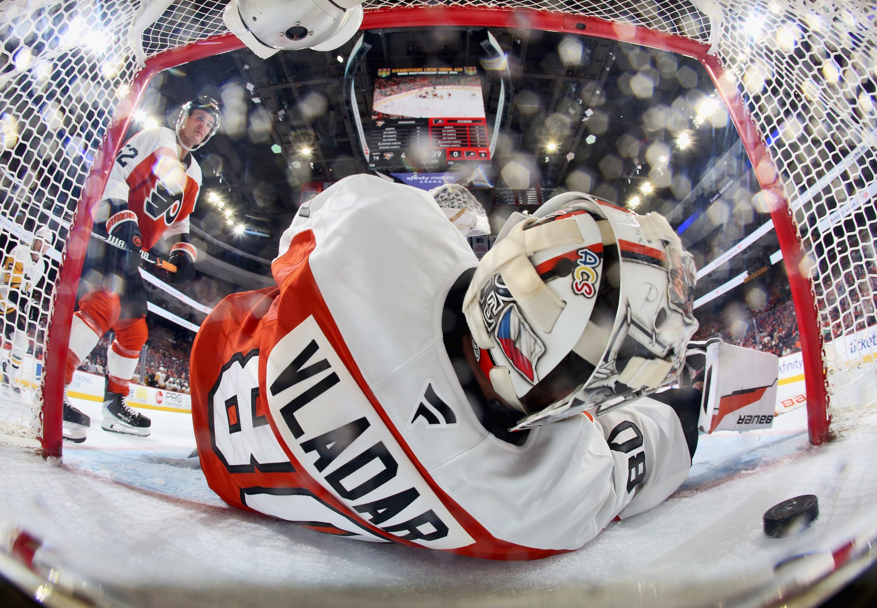 Dan Vladar of the Philadelphia Flyers surrenders a goal during the second period against the Pittsburgh Penguins as Christian Dvorak #22 looks on. Half his body is in the goal, and the puck is visible at lower right.