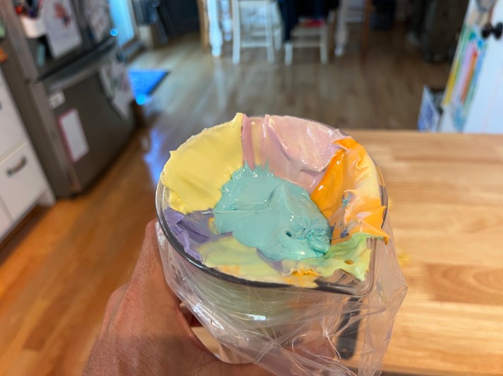 Six blobs of colored icing scooped into a piping bag pulled over a plastic pitcher.